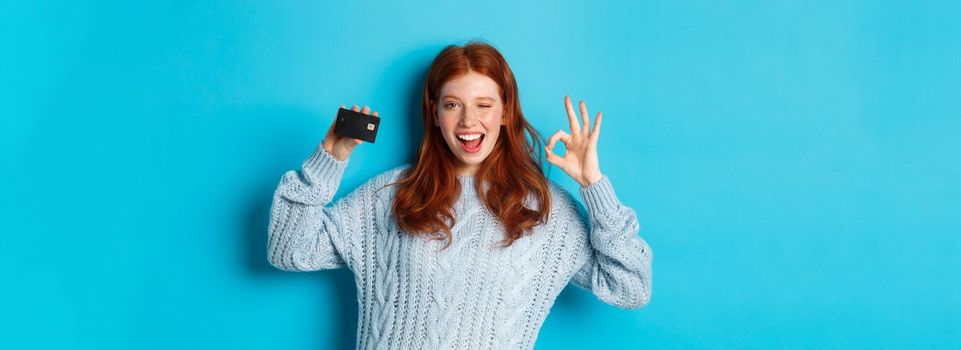 Happy redhead girl in sweater showing credit card and okay sign, recommending bank offer, standing over blue background.