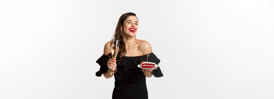 Celebration and party concept. Fashionable woman holding birthday cake with candle and drinking champagne, laughing happy, standing over white background.