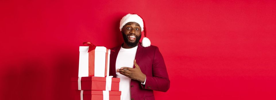 Christmas, New Year and shopping concept. Cheerful Black man secret santa holding xmas presents and smiling excited, bring gifts, standing against red background.