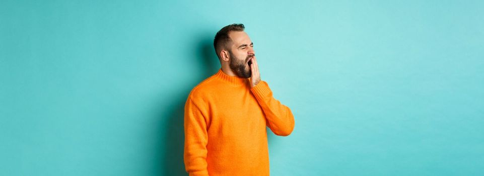 Photo of tired bearded man yawning, cover mouth with hand, standing sleepy against light blue background.