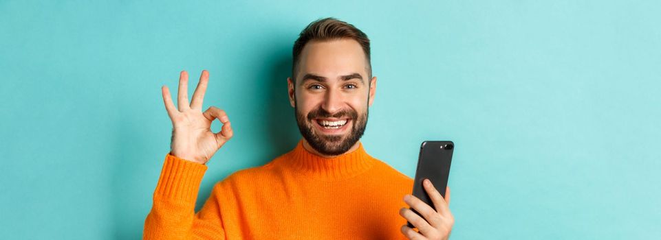 Close-up of bearded man holding smartphone and showing okay sign, recommending mobile app, standing satisfied over turquoise background.