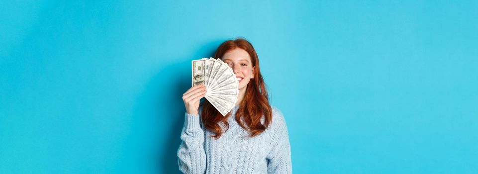 Delighted redhead woman showing money near face, smiling satisfied, standing ready for shopping over blue background.