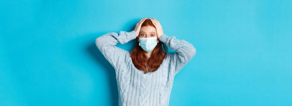 Winter, covid-19 and social distancing concept. Troubled redhead girl in face mask staring distressed, holding hands on head in panic, standing over blue background.