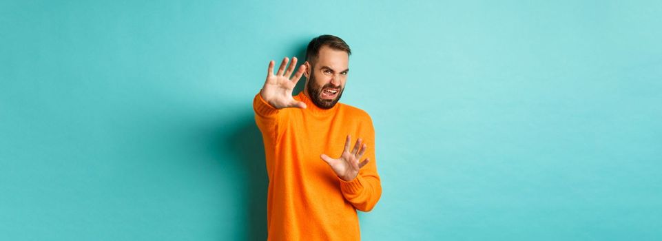 Disgusted man cringe and extending hands in protection, begging for mercy, looking at something with aversion, standing over light blue background.