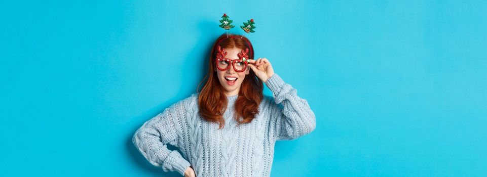 Christmas party and celebration concept. Cute redhead teen girl celebrating New Year, wearing xmas tree headband and funny glasses, looking left amused, blue background.