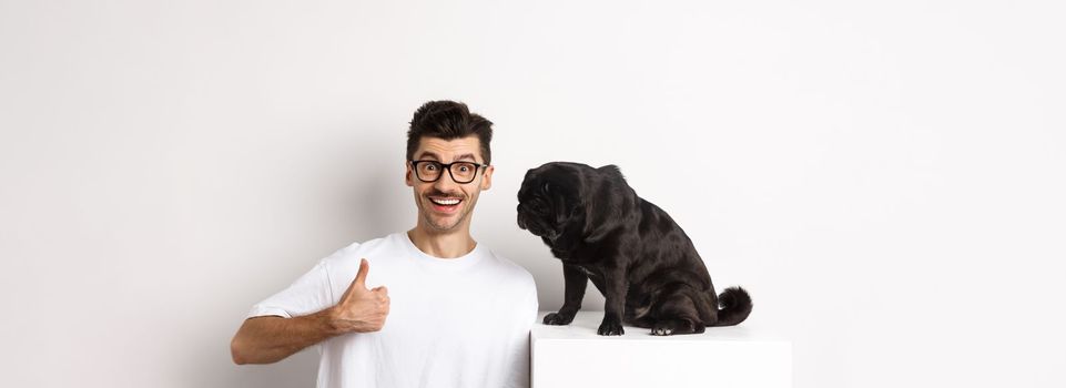 Image of happy and satisfied young man sitting near pug dog and showing thumbs-up, smiling and praising good product, white background.