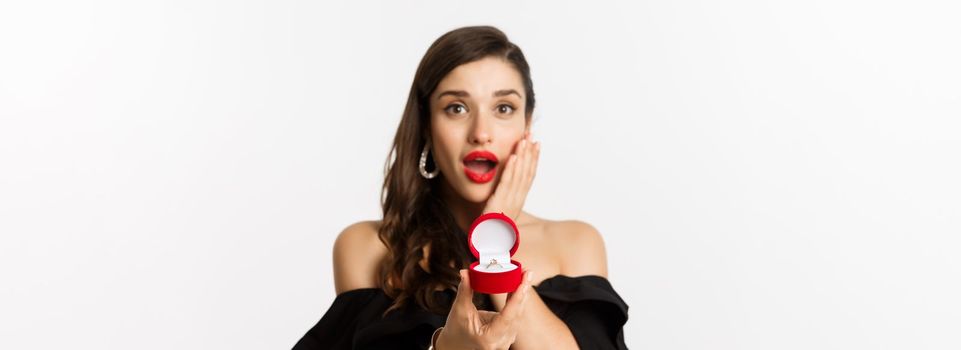 Beautiful woman in black dress making proposal, showing engagement ring and looking excited, standing over white background.