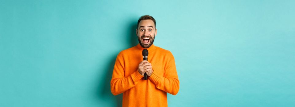 Happy adult man singing karaoke, holding microphone and looking at camera, standing in orange sweater against turquoise background.