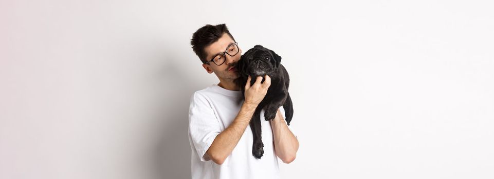 Handsome young man pet his cute black dog, scratching pug while holding animal on shoulder, standing over white background.
