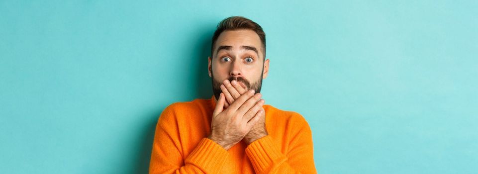 Close-up of shocked caucasian man covering his lips and mouth, staring astounded at camera, standing against turquoise background.