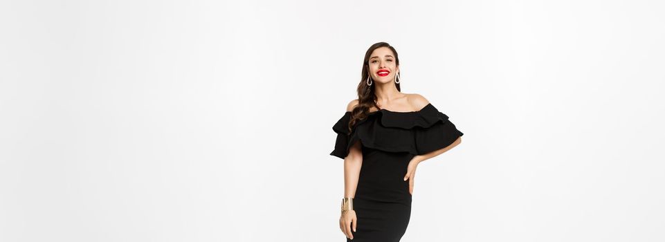 Beauty and fashion concept. Full length shot of beautiful brunette woman wearing luxury black dress and heels on party, smiling with red lips, standing over white background.