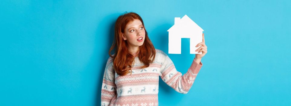 Real estate concept. Image of cute redhead girl looking curious at paper house model, thinking of buying property, standing in sweater against blue background.