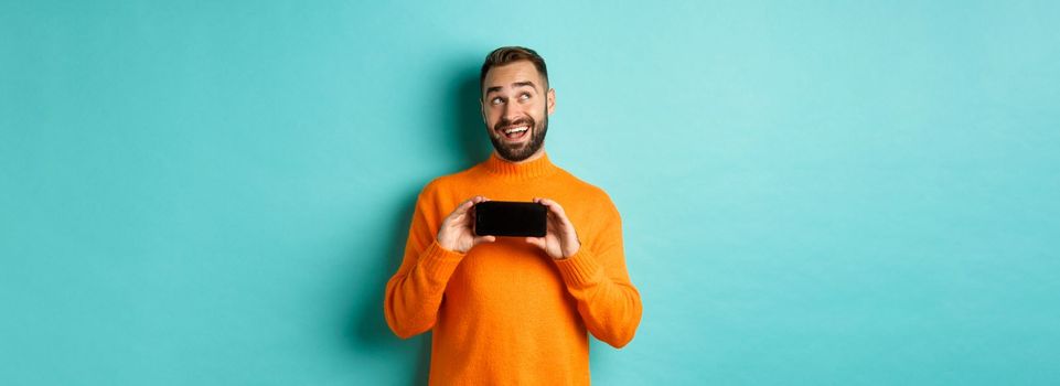 Online shopping. Young man thinking and showing smartphone screen, looking dreamy at upper left corner, standing over light blue background.