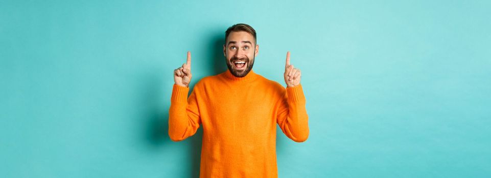 Happy caucasian man showing winter holidays promo offer, pointing fingers up, demonstrate advertisement, standing in sweater against turquoise background.