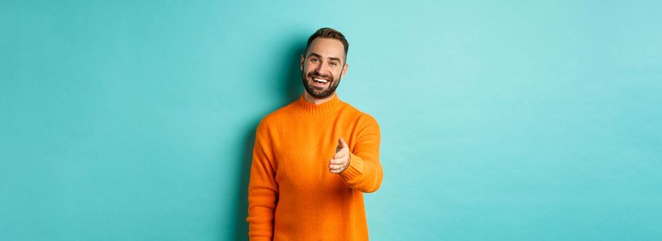 Friendly nice guy extend hand for handshake, greeting you and smiling, saying hello, standing over light blue background.