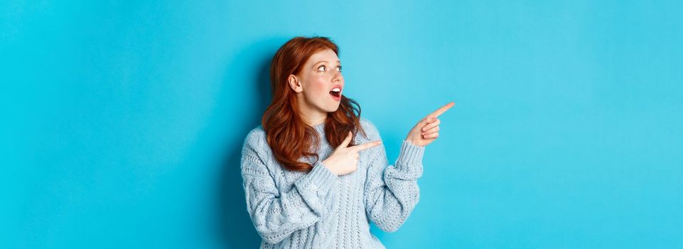 Winter holidays and people concept. Amazed redhead girl checking out promo offer, pointing and looking left at copy space, standing against blue background.