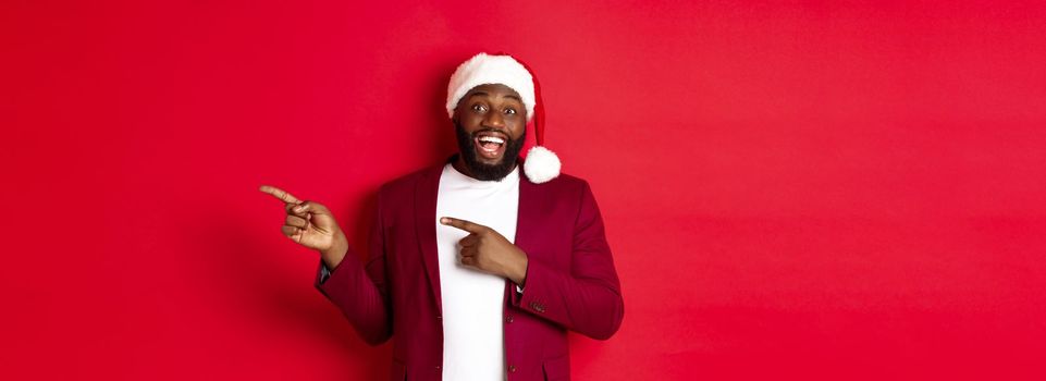 Christmas, party and holidays concept. Cheerful Black man smiling, pointing fingers right and showing advertisement, standing over red background.