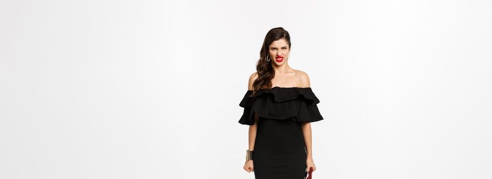 Beauty and fashion concept. Full length of angry woman in black party dress and high heels, express disdain and grimacing at camera, mad at person, white background.