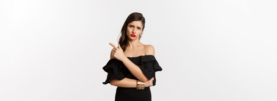 Fashion and beauty. Skeptical glamour woman with red lips, black dress, pointing finger right at something lame and boring, standing over white background.