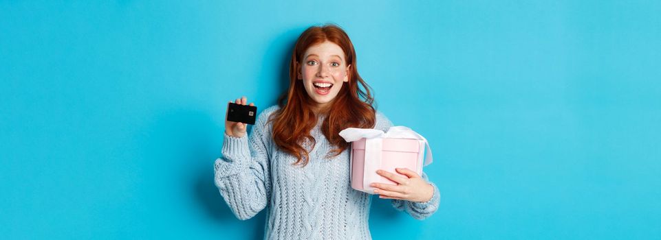 Winter holidays promo offer concept. Cheerful redhead woman holding Christmas gift and credit card, staring at camera amazed, standing over blue background.