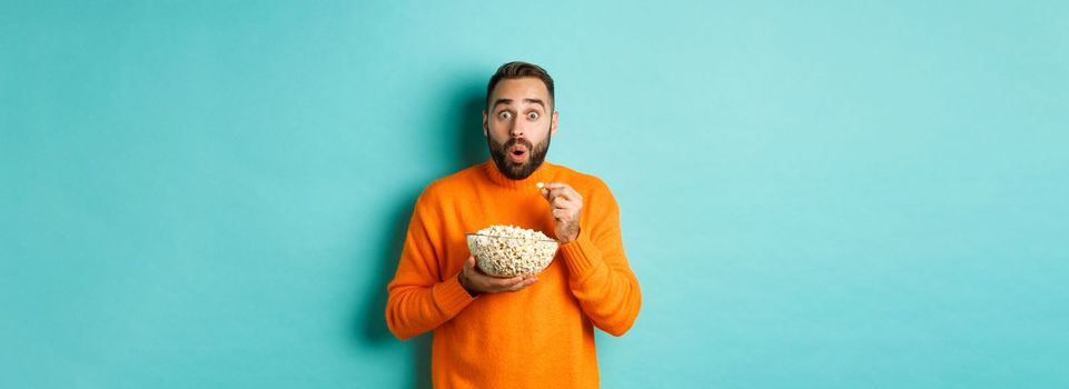 Excited young man watching interesting movie on tv screen, eating popcorn and looking amazed, blue background.