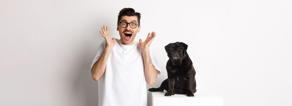 Ecstatic young man looking with excitement and rejoice, standing near cute black pug, staring at camera happy, standing over white background.