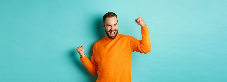 Winner. Excited man triumphing, making fist pump and saying yes, triumphing and looking satisfied, achieve goal, standing over light blue background.