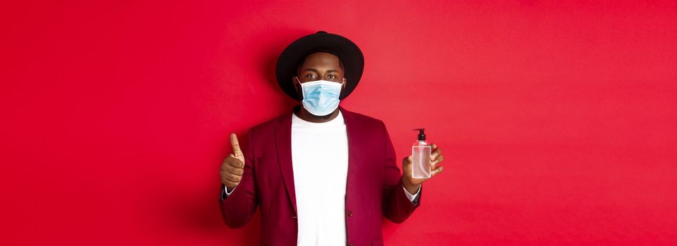 Covid-19, quarantine and holidays concept. Young african american man in medical mask, showing hand sanitizer and thumb up, recommending to use antiseptic.