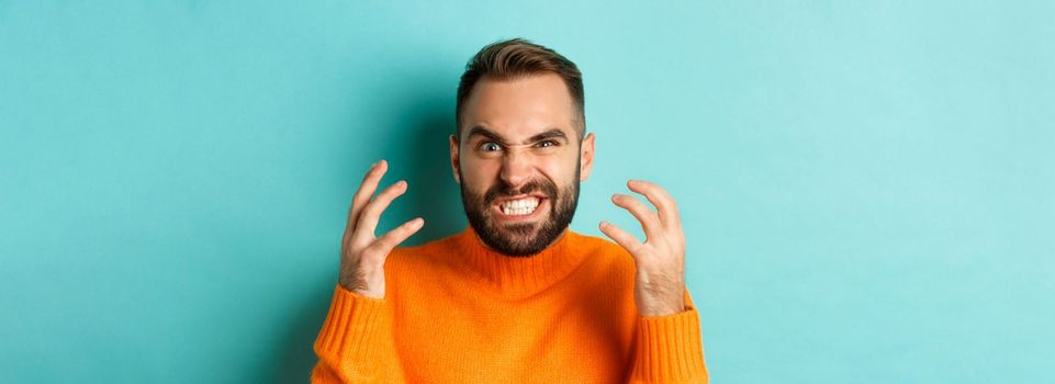 Close-up of angry caucasian man looking mad, grimacing and shaking hands, having break down, standing over light blue background.