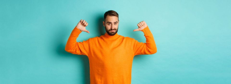 Confident attractive guy pointing at himself, show-off or brag, standing over light blue background.