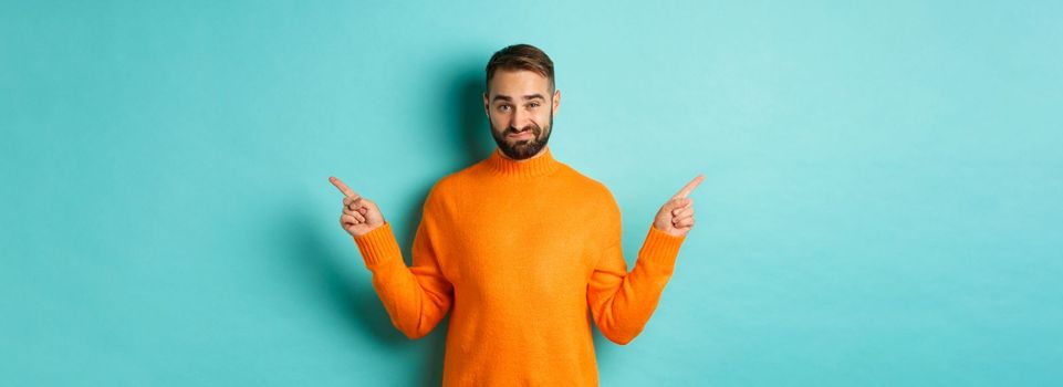 Doubtful and indecisive man pointing fingers sideways, showing different sides, two choices, looking skeptical, standing over light blue background.