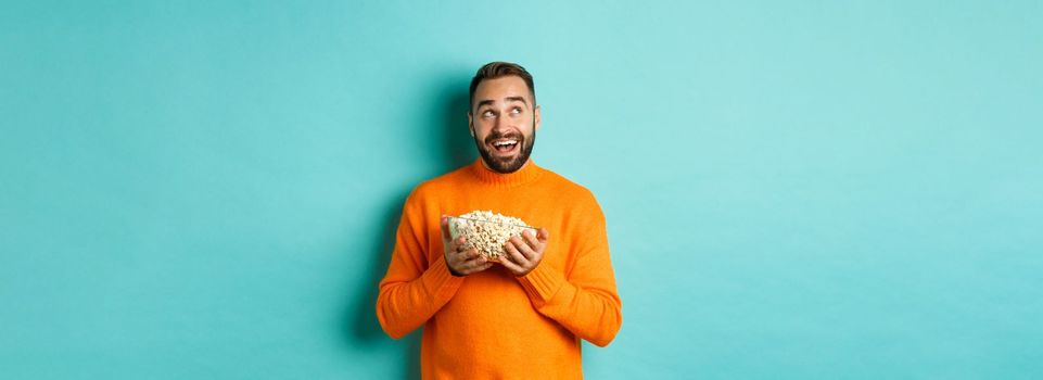 Handsome young man in orange sweater, looking thoughtful at upper left corner, holding popcorn, picking movie, blue background.