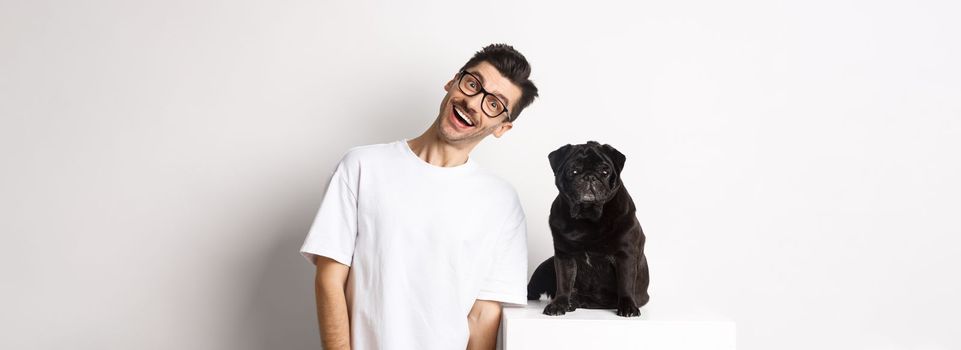 Image of handsome young man standing near cute black pug and smiling. Dog owner spending time with his pet, staring at camera happy, white background.