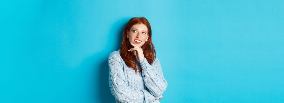 Thoughtful teenage girl with red hair, looking upper right corner logo and thinking, imaging something, standing over blue background.