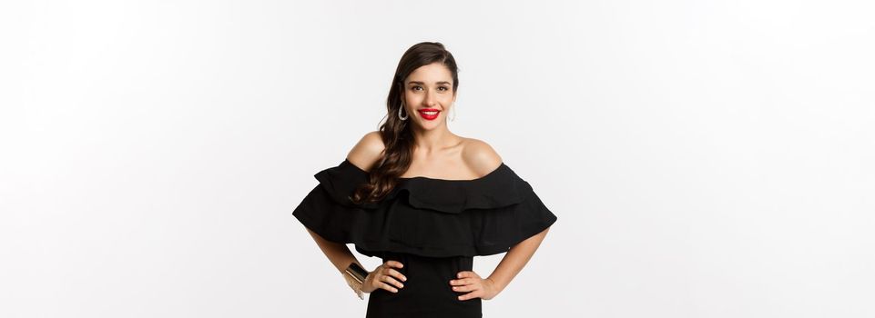 Waist-up shot of beautiful woman with red lipstick, wearing black dress and smiling pleased, standing over white background.