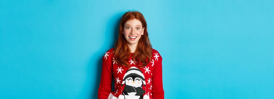 Winter holidays and Christmas Eve concept. Excited redhead girl in xmas sweater looking surprised at camera, standing against blue background.