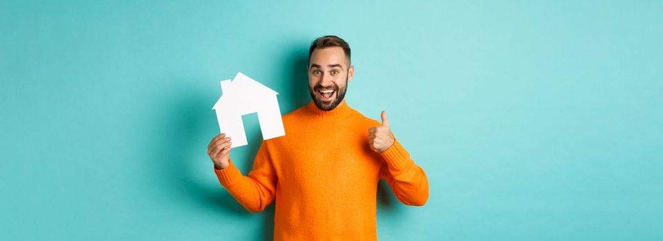 Real estate concept. Satisfied man recommending agency, showing thumb up and house maket, standing over blue background.