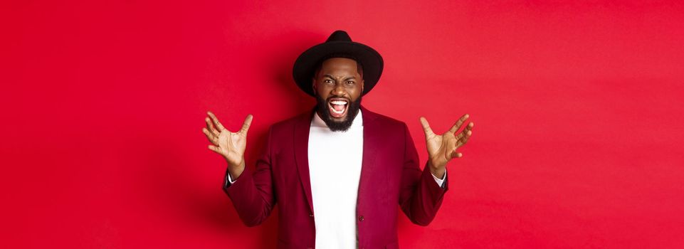 Angry Black man screaming and shaking hands with hatred, losing temper, standing outraged against red background.