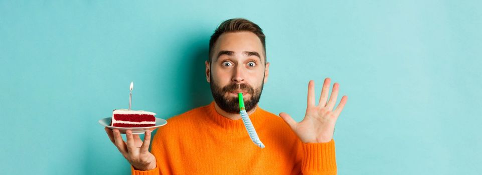 Close-up of funny adult man celebrating his birthday, holding bday cake with candle, blowing party wistle and rejoicing, standing over light blue background.