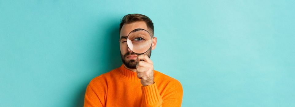 Funny man looking through magnifying glass, searching or investigating something, standing in orange sweater against turquoise background.