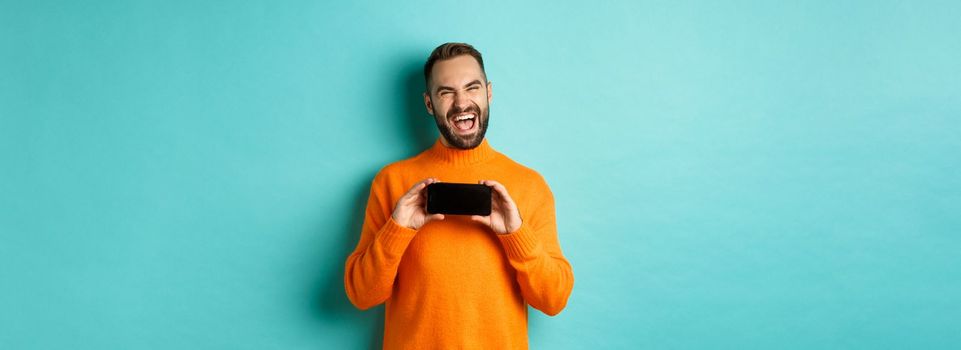 Photo of happy adult man laughing and showing funny thing on mobile phone screen, standing in orange sweater over turquoise background.