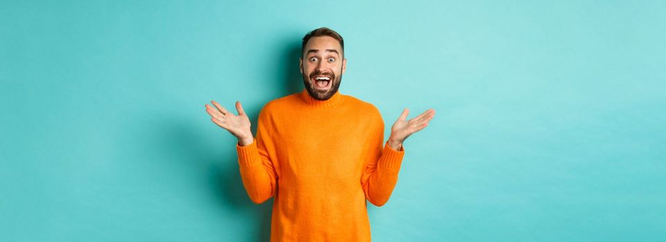 Surprised man raising hands up, looking at something amazing, standing over turquoise background in winter sweater.