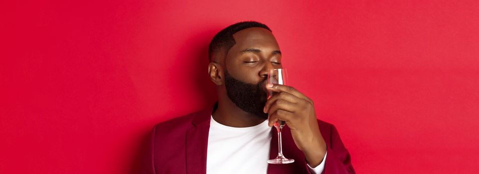 Drunk african american guy kissing glass with champagne, celebrating new year, enjoying party, standing over red background.