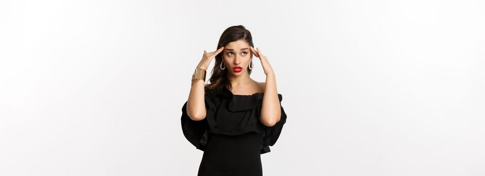 Fashion and beauty. Annoyed and tired woman in black dress, touching head and roll eyes bothered, standing distressed against white background.