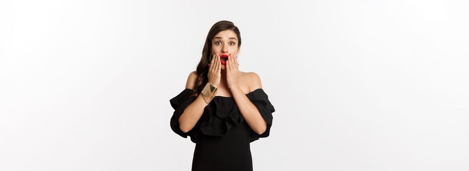 Fashion and beauty. Image of attractive female model in black dress reacting to announcement, looking amazed at camera, standing surprised over white background.