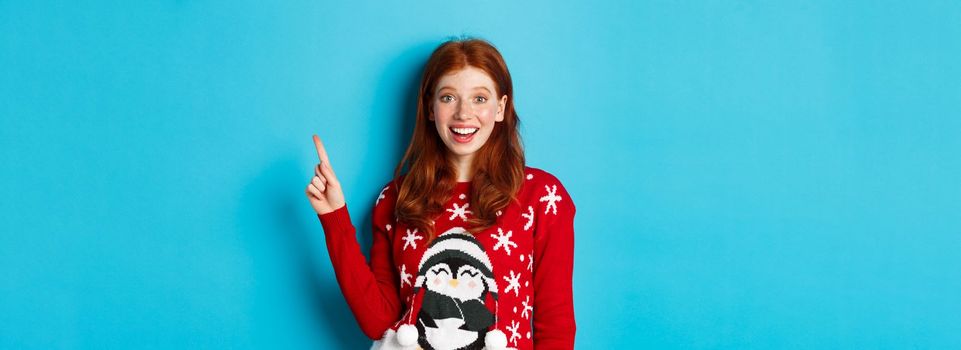 Merry Christmas. Cheerful redhead girl in xmas sweater, pointing finger at upper right corner and smiling excited, showing new year promo.