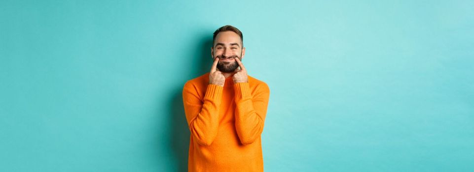 Image of bearded man stretching lips in happy smile, faking happiness, standing over light blue background. Copy space