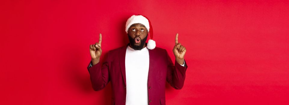 Christmas, party and holidays concept. Impressed Black man in santa hat pointing fingers up, saying wow and staring at camera, checking out promo offer, red background.
