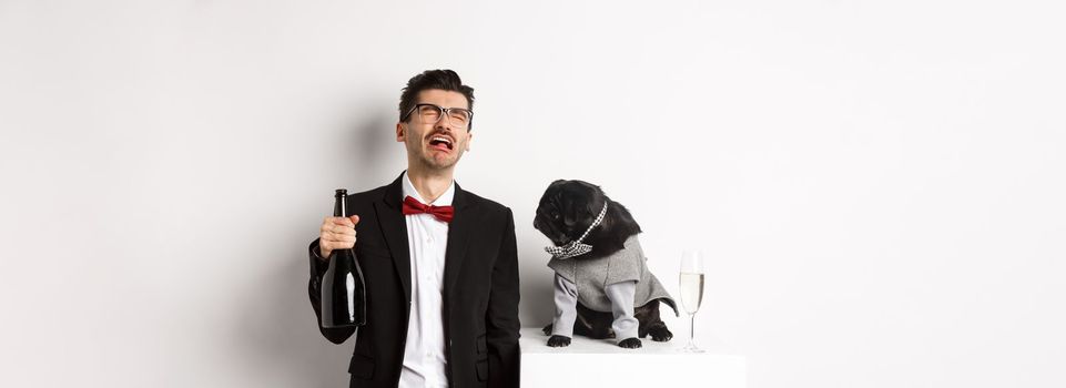 Sad man crying and drinking champagne from bottle while pug in cute party costume staring confused, standing over white background.