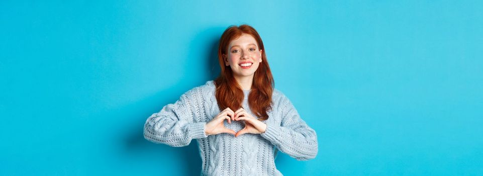 Cute redhead girl in sweater showing heart sign, I love you gesture, smiling at camera, standing against blue background.
